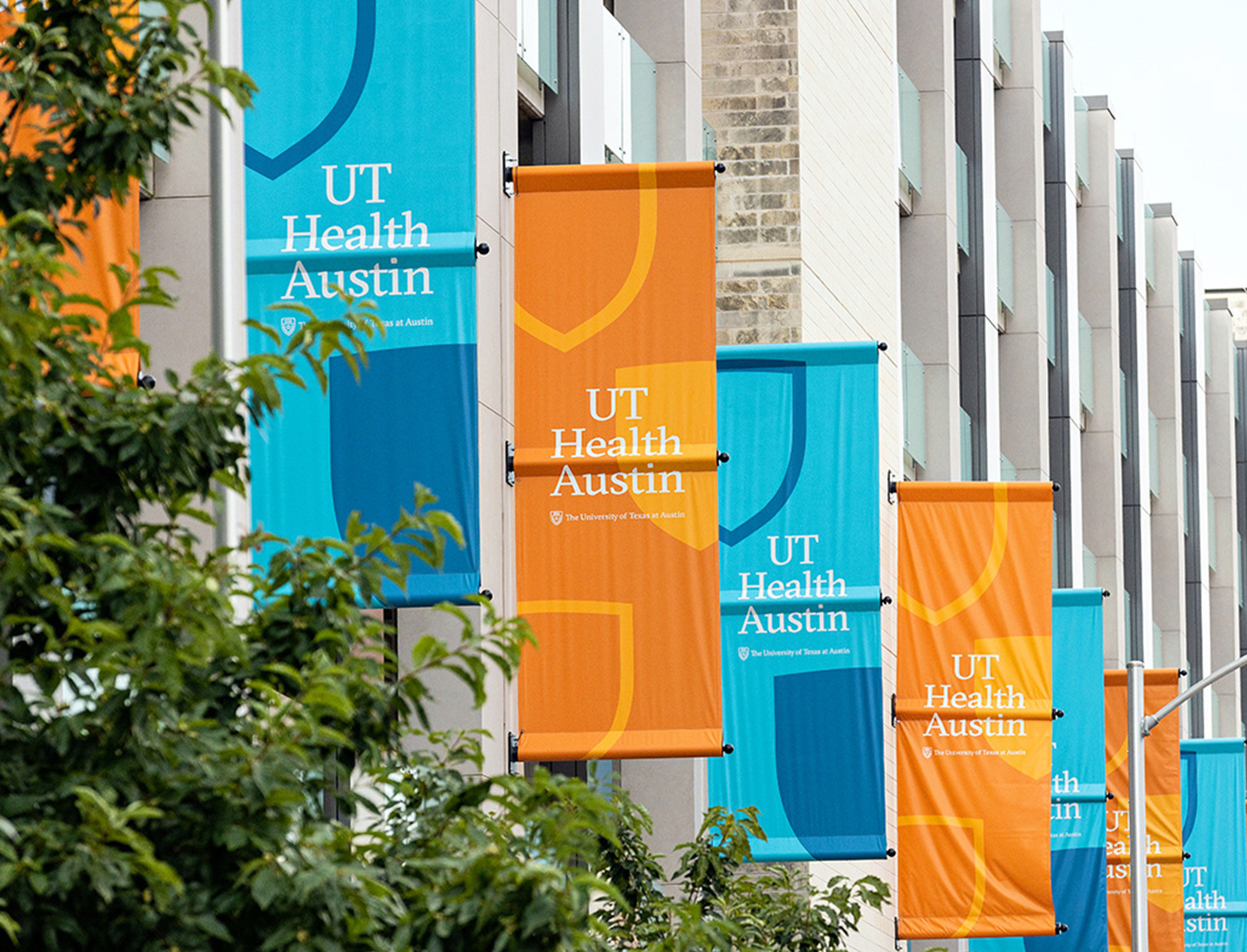 UT Health Austin building's west façade with UT Health Austin flags in alternating colors of teal and burnt orange.