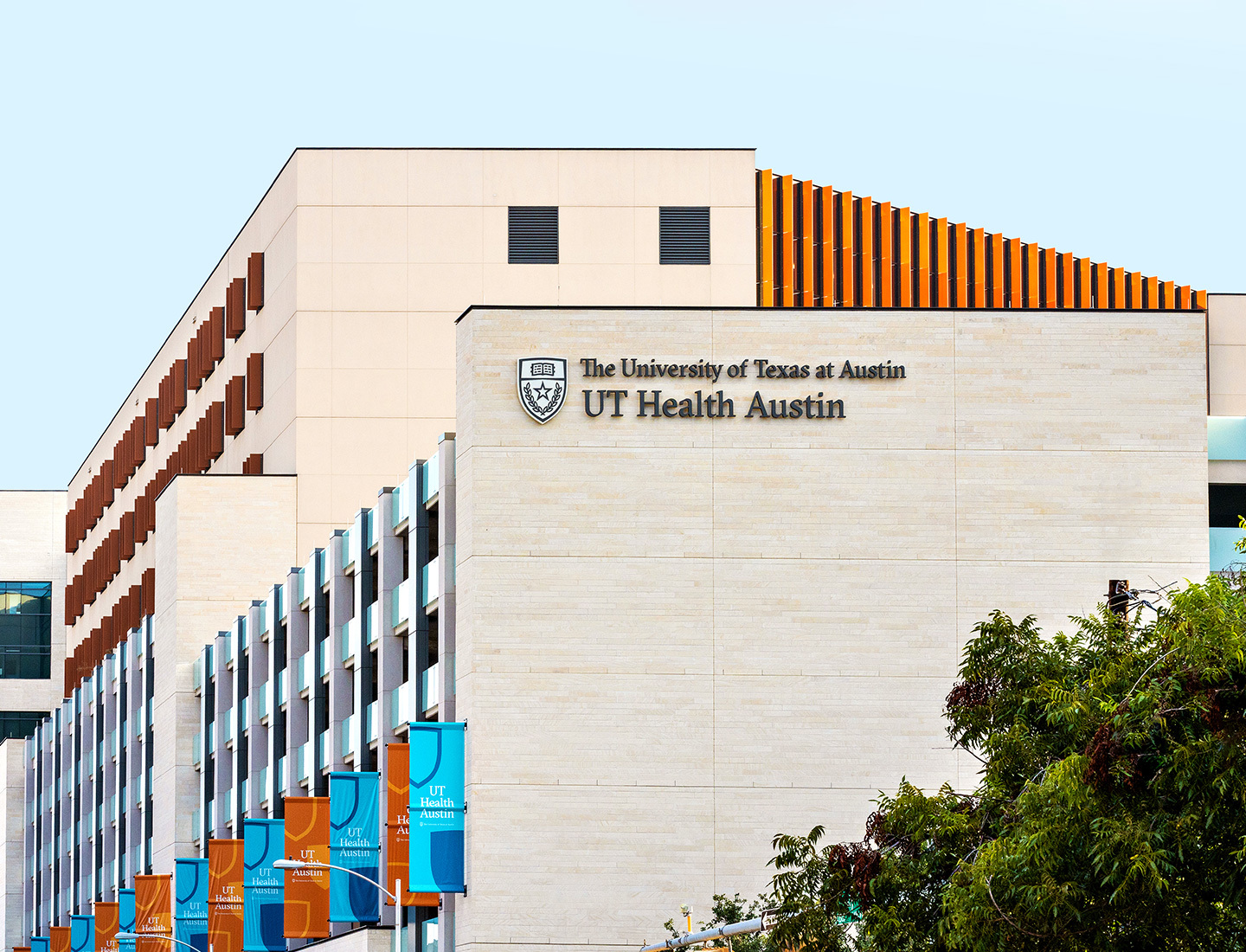 UT Health Austin sign on the south side of the Health Transformation Building as seen from the street with alternating flags of teal and burnt orange on the west side.